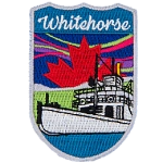 Whitehorse is stitched above a white river boat. There is a red maple leaf in the sky with the northern lights.