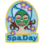 A woman with a green face mask, a towel wrapped around her head, and flowers around her. The words Spa Day are stitched below.