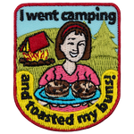 A woman holds two burnt buns to the viewer with a campsite behind her.
