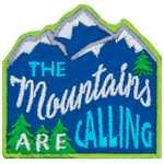 The words The Mountains Are Calling are stitched across a mountain range with pine trees. 