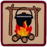 A black iron pot hangs from a high bar suspended on forks which are all made from branches. The pot simmers over a roaring campfire.
