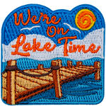 A blue, sunny sky above white puffy clouds. A wooden dock extends into a calm lake. We're On Lake Time, sits in the sky.