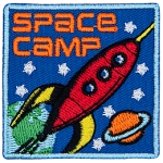 A red rocket blasts through space past Earth and Saturn. The words Space Camp are in the top left corner.