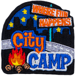 The words Where Fun Happens are on a signpost. Below them, overtop a road are the words City Camp and a campfire. The back is a night skyline.