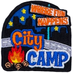 The words Where Fun Happens are on a signpost. Below them, overtop a road are the words City Camp and a campfire. The back is a night skyline.