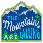 The words The Mountains Are Calling are stitched across a mountain range with pine trees. 