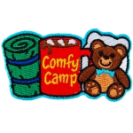 From left to right is a sleeping bag, a mug of coco with the words 'Comfy Camping' on the side, and a teddy bear with a pillow behind it.