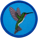 A hummingbird with wings spread wide open faces the right edge of this round circle badge.