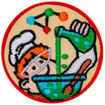 A ginger boy with a chef hat and a lab coat pours a beaker of bubbling green liquid into a blue bowl. A complex molecule floats above the chef's head.