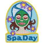 A woman with a green face mask, a towel wrapped around her head, and flowers around her. The words Spa Day are stitched below.