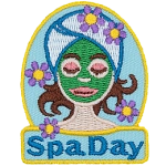 A woman with a green face mask, a towel wrapped around her head, and flowers around her. The words Spa Day are stitched below.