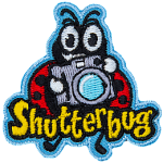 A ladybug holds up a retro camera. The word Shutterbug is stitched across the bottom.  Photography.