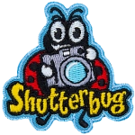 A ladybug holds up a retro camera. The word Shutterbug is stitched across the bottom.  Photography.