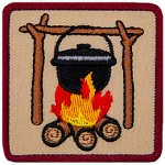 A black iron pot hangs from a high bar suspended on forks which are all made from branches. The pot simmers over a roaring campfire.