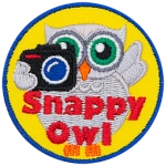 A grey owl holds a camera with his left-wing while the other is up in the air. The words Snappy Owl are written in red near the bottom of the crest.