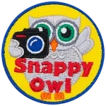A grey owl holds a camera with his left-wing while the other is up in the air. The words Snappy Owl are written in red near the bottom of the crest.