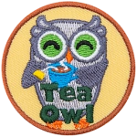 A grey owl smiles and holds a steaming cup of tea with the tea bag still in it. At the bottom of this crest are the words Tea Owl.