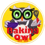 An owl stands in a white cooking apron and holds a bowl in its left wing and a whisk in its right. The words Baking Owl rest at the bottom center of the crest.