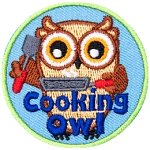 A brown owl stands holding a spatula in its left wing and a steaming frying pan in its right. The words Cooking Owl rest at the bottom center of the crest.