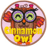 A brown owl with purple glasses with green lenses. The words Cinnamon Owl are stitched across the bottom.