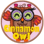 A brown owl with purple glasses with green lenses. The words Cinnamon Owl are stitched across the bottom.