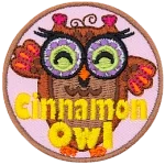 A brown owl with purple glasses with green lenses. The words Cinnamon Owl are stitched across the bottom.