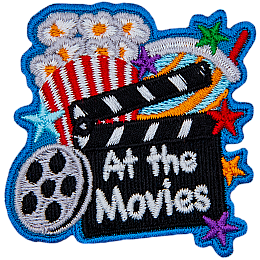 A clapperboard with the words At The Movie rests against a popcorn bucket. A roll of film is next to it.