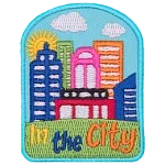 A sunny cityscape sits on top of a green hill. The words In The City are embroidered below.