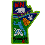 This crest is shaped like the province of Manitoba. Decorating it are the initials MB, a polar bear, the aurora Borealis, a snowflake, two loonie coins, a lake, a pair of slippers, and the capital Winnipeg.