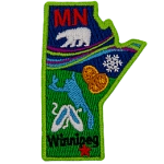 This crest is shaped like the province of Manitoba. Decorating it are the initials MB, a polar bear, the aurora Borealis, a snowflake, two loonie coins, a lake, a pair of slippers, and the capital Winnipeg.