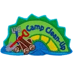 A green and yellow sea serpent with the words Camp Clean-Up on it and a pile of wood, a shove and a rake next to it.